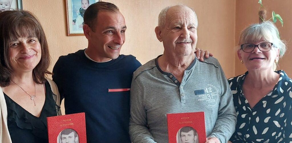 Quatre adultes souriants entourent un homme âgé tenant un livre rouge avec une photo en noir et blanc sur la couverture. Une scène chaleureuse de transmission et de mémoire familiale.